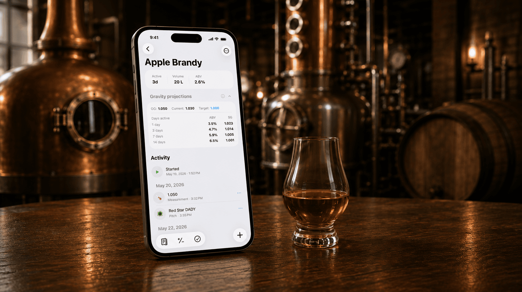 A phone displaying Mashpad's ferment-tracking screen on a wood table in a craft distillery, with copper stills and a glencairn of amber spirit